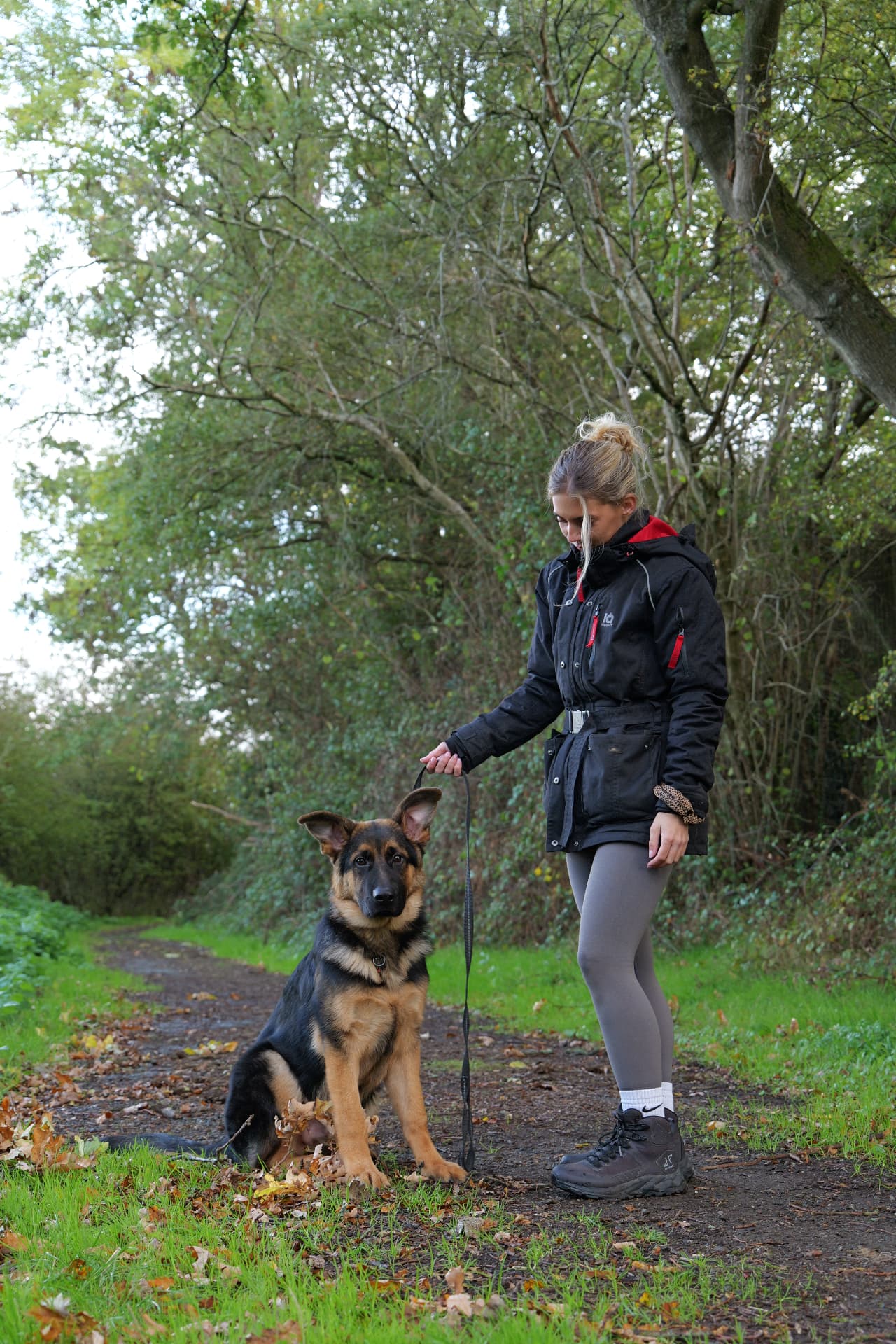 Holly with a German Shepherd puppy sitting calmly on a woodland path